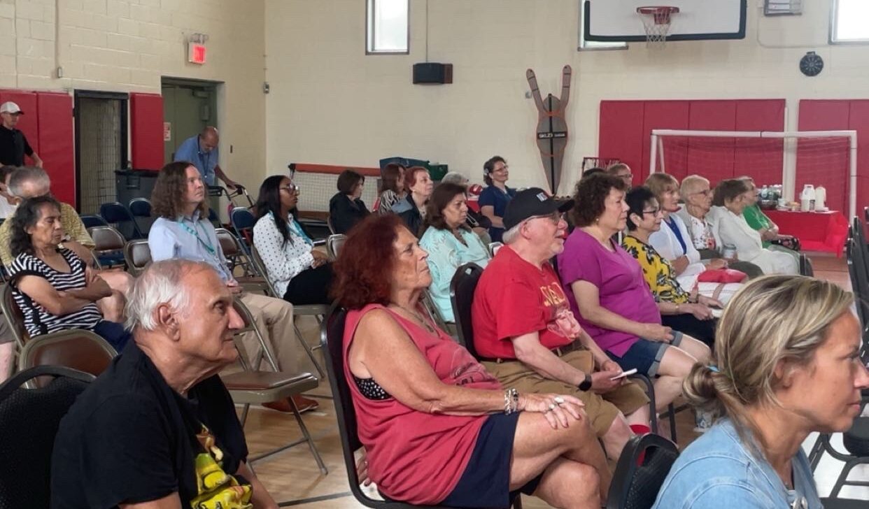 People sit on chairs in a gymnasium in New York, attentively watching a Model Film Screening focused on Aging in Community. The space features a basketball hoop, high windows, and wooden floors.