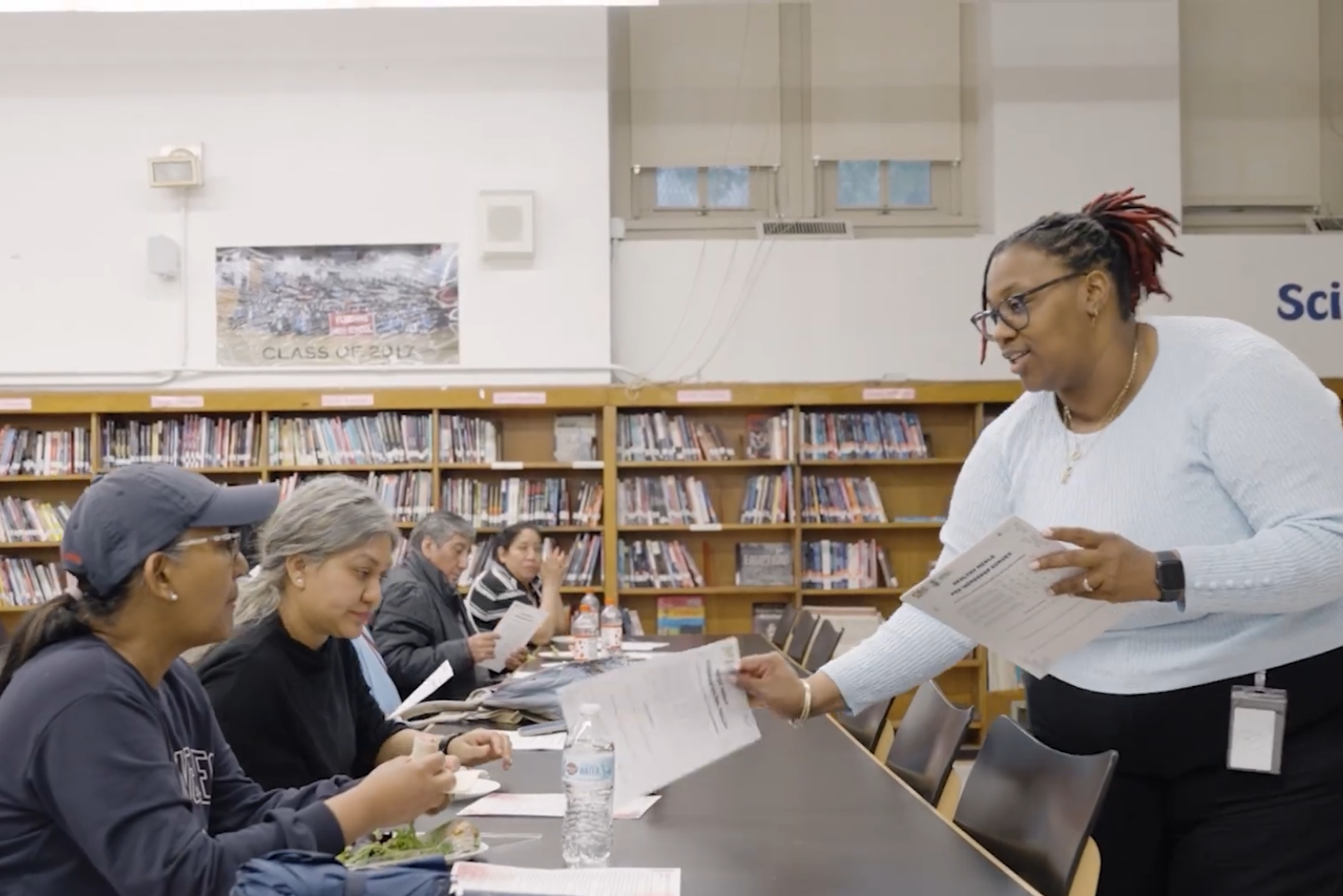 A woman hands papers to two seated people in a library, with bookshelves and a "Science" sign visible in the background—part of the Healthy Meals Project by Commonpoint Queens.