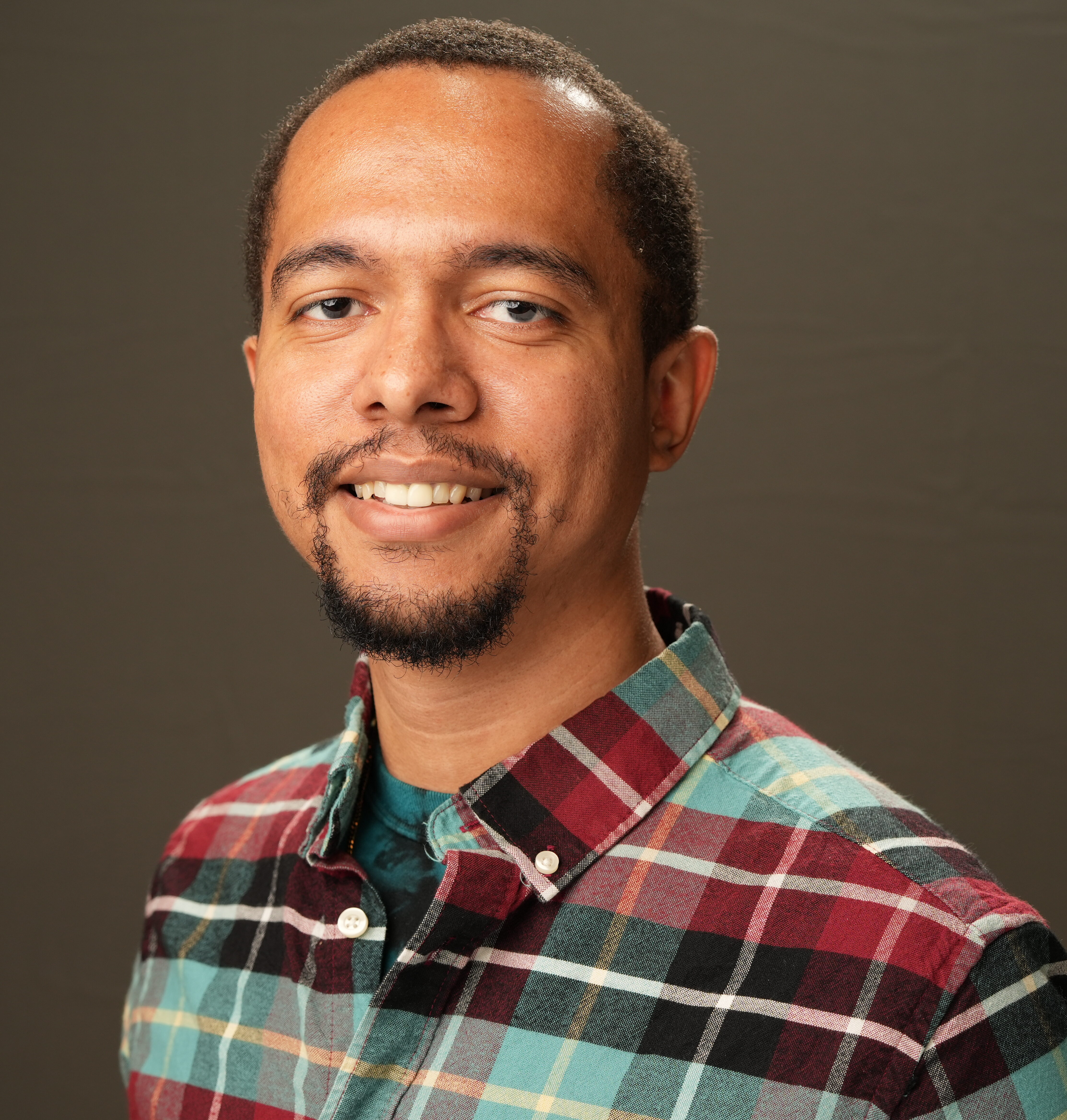 Charles Witherspoon, a man with short hair and a beard, wearing a plaid shirt, smiles at the camera against a plain dark background.