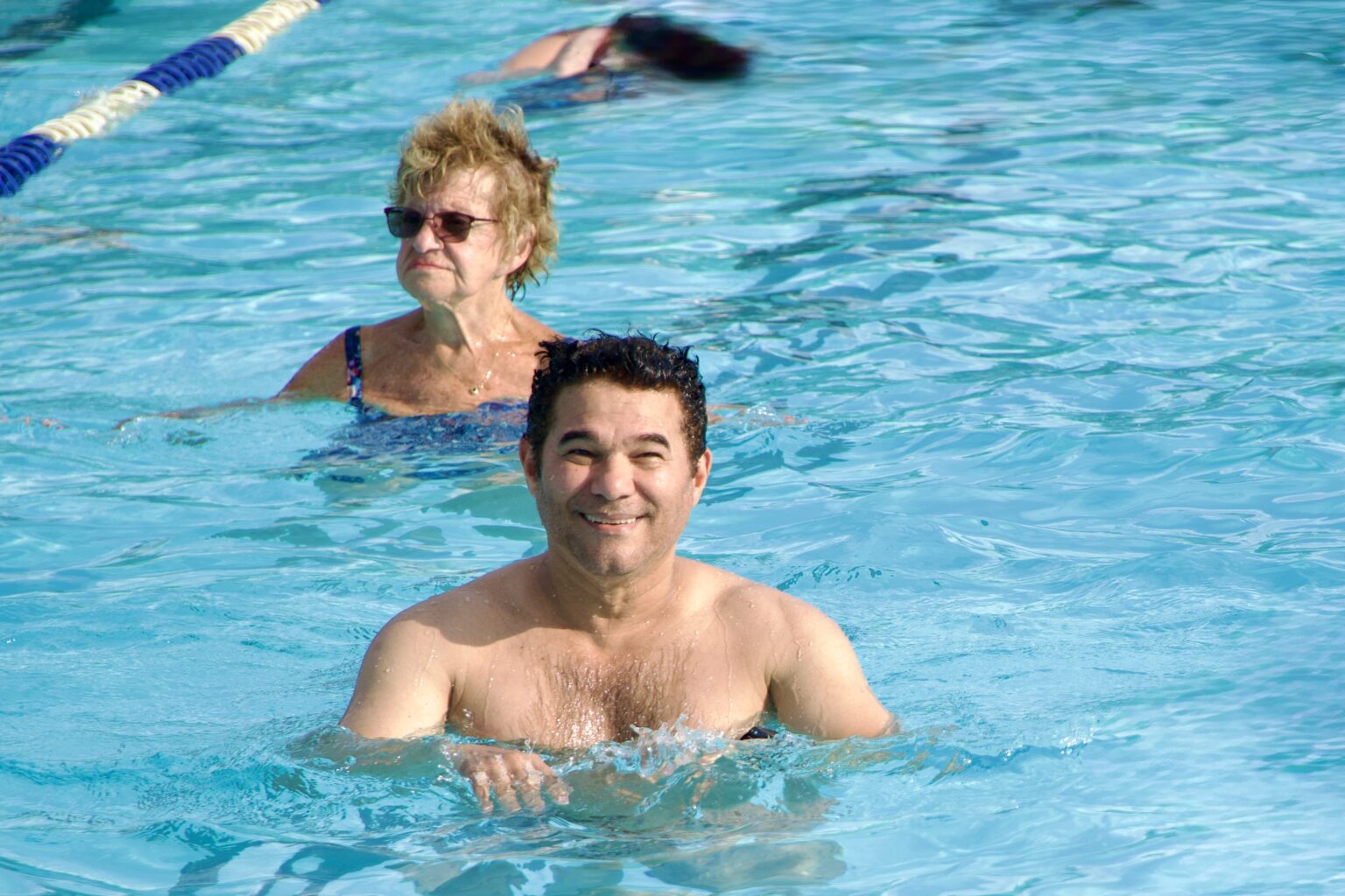 A man and an older woman are swimming in an outdoor pool, with another person swimming in the background.