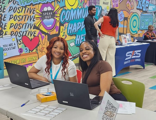 Two women sit at a table with laptops, smiling at the camera. Behind them is a colorful mural and a few people standing near informational booths.