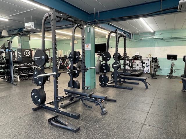 A gym with squat racks, barbells, weight plates, benches, and exercise equipment on a rubber floor. The room has teal beams and fluorescent lighting.