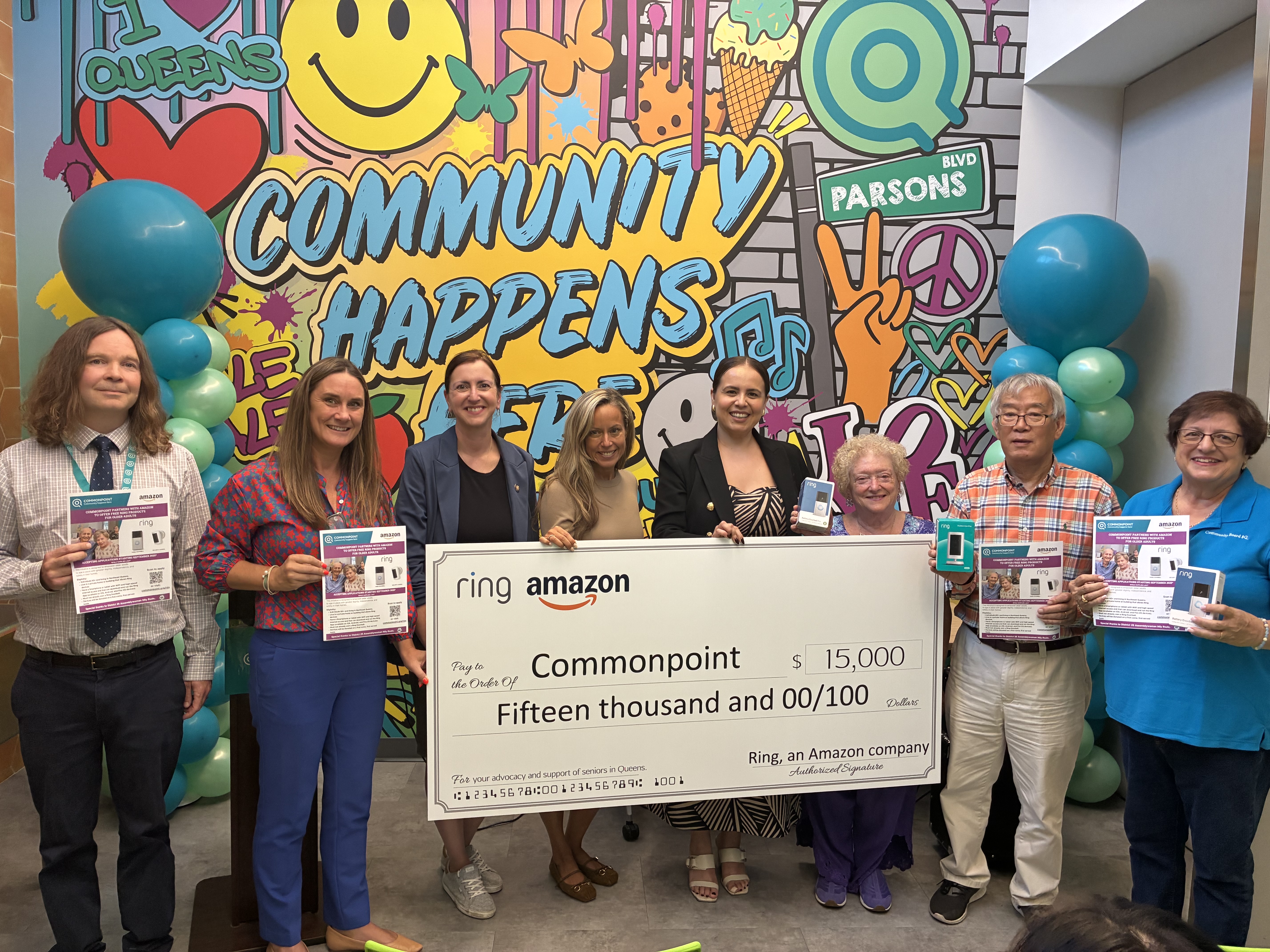 A group stands in front of a colorful mural holding a $15,000 check from Ring and Amazon to Commonpoint, highlighting their commitment to community and home security. Some people are also holding flyers.
