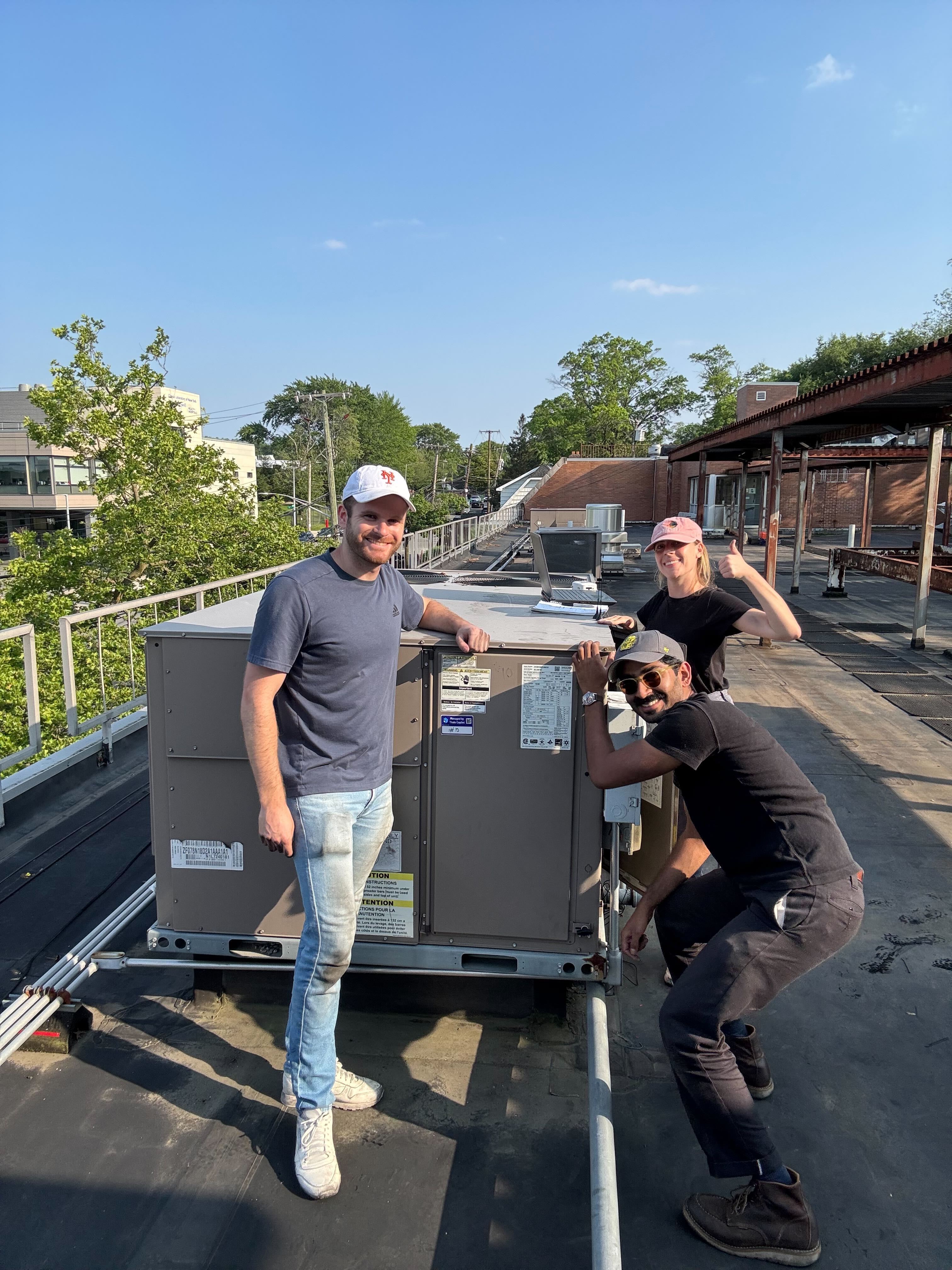 Three people stand and pose next to rooftop HVAC equipment on a sunny day at Commonpoint NYC Community Center, with trees and buildings visible in the background.