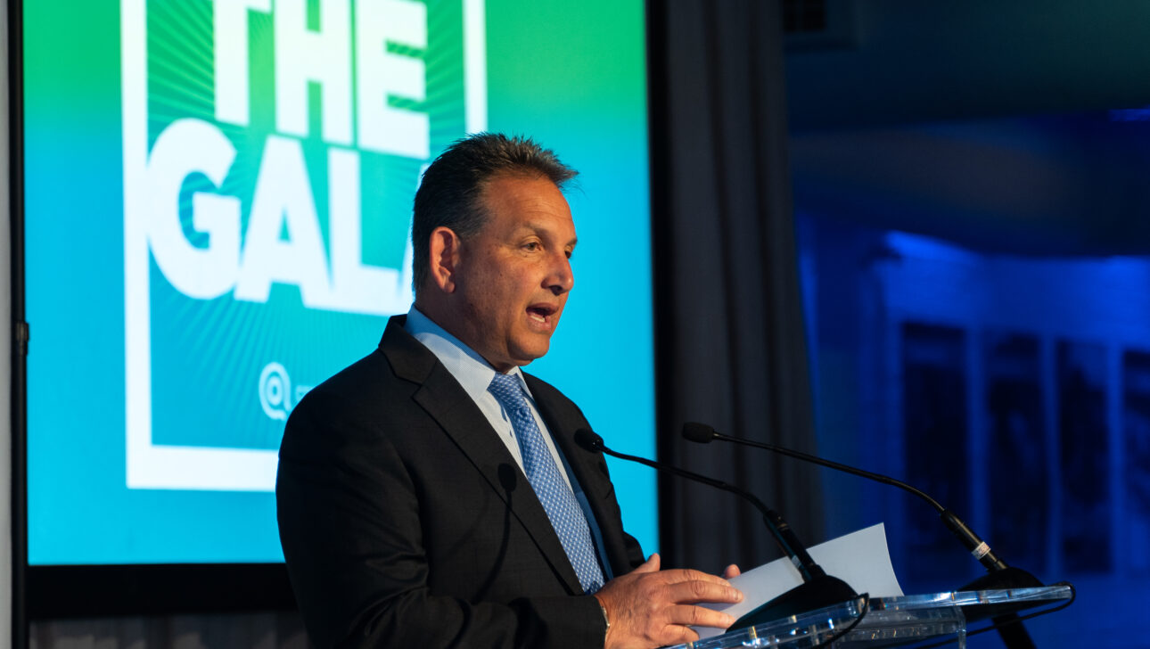 A man in a suit speaks at a podium with microphones, addressing the audience at the Commonpoint Gala, as "THE GALA" is displayed in bold letters on a large screen behind him.