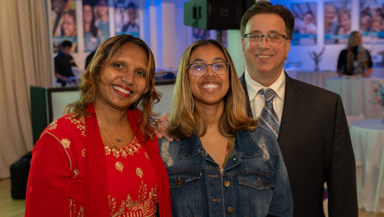 Three people stand together at the Commonpoint Gala; the woman on the left wears a red dress, the woman in the center sports glasses and a denim jacket, and the man on the right is dressed in a suit and tie.