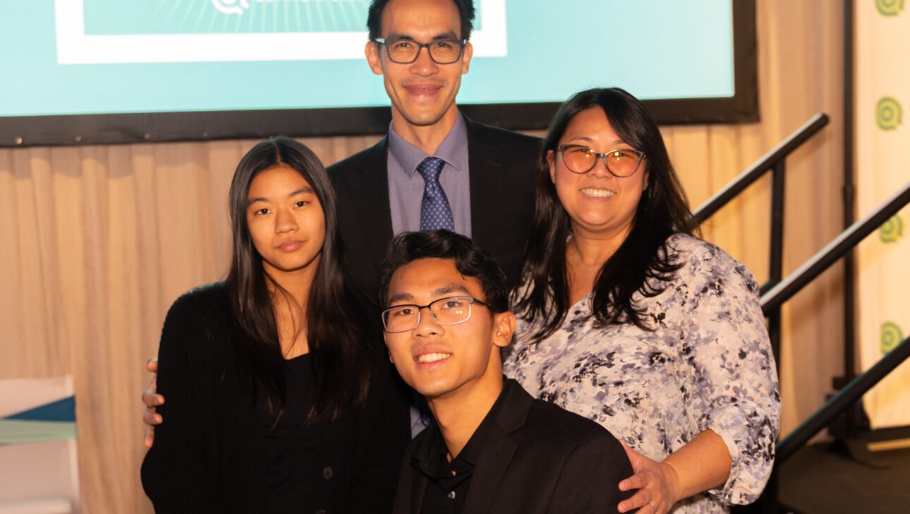 Four people pose and smile in front of a presentation screen at the Commonpoint Gala. Three are standing, one is seated. All are dressed in semi-formal attire at this indoor event.
