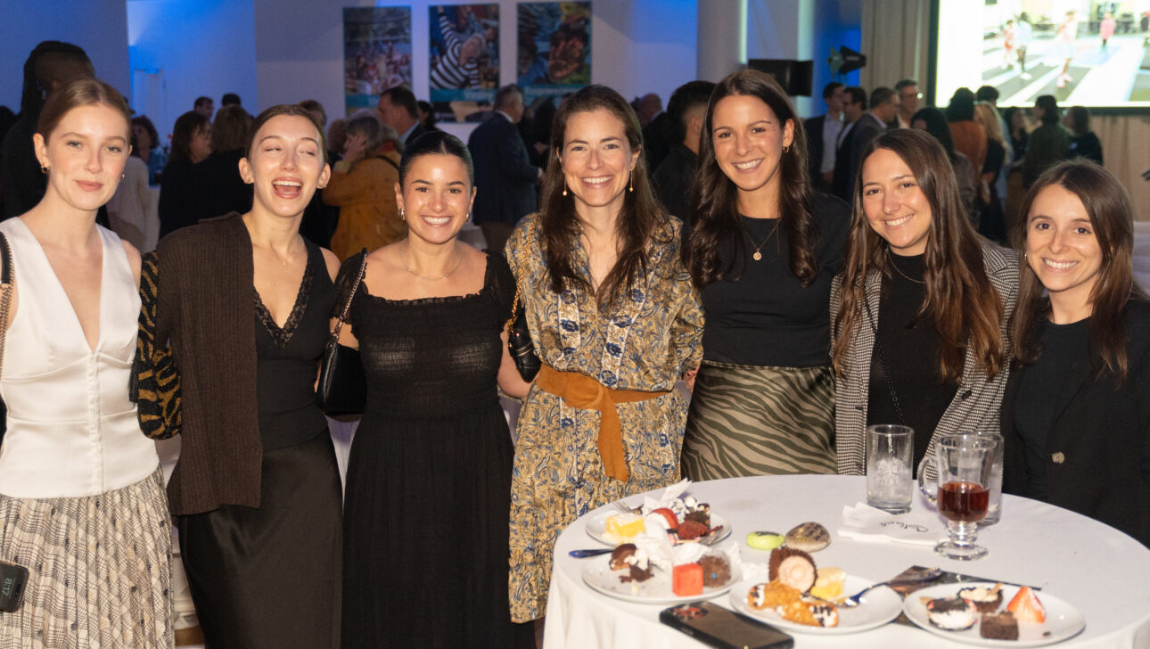 Seven women stand together and smile for a group photo at the Commonpoint Gala, gathered around a tall table with plates of desserts and drinks at this indoor event.
