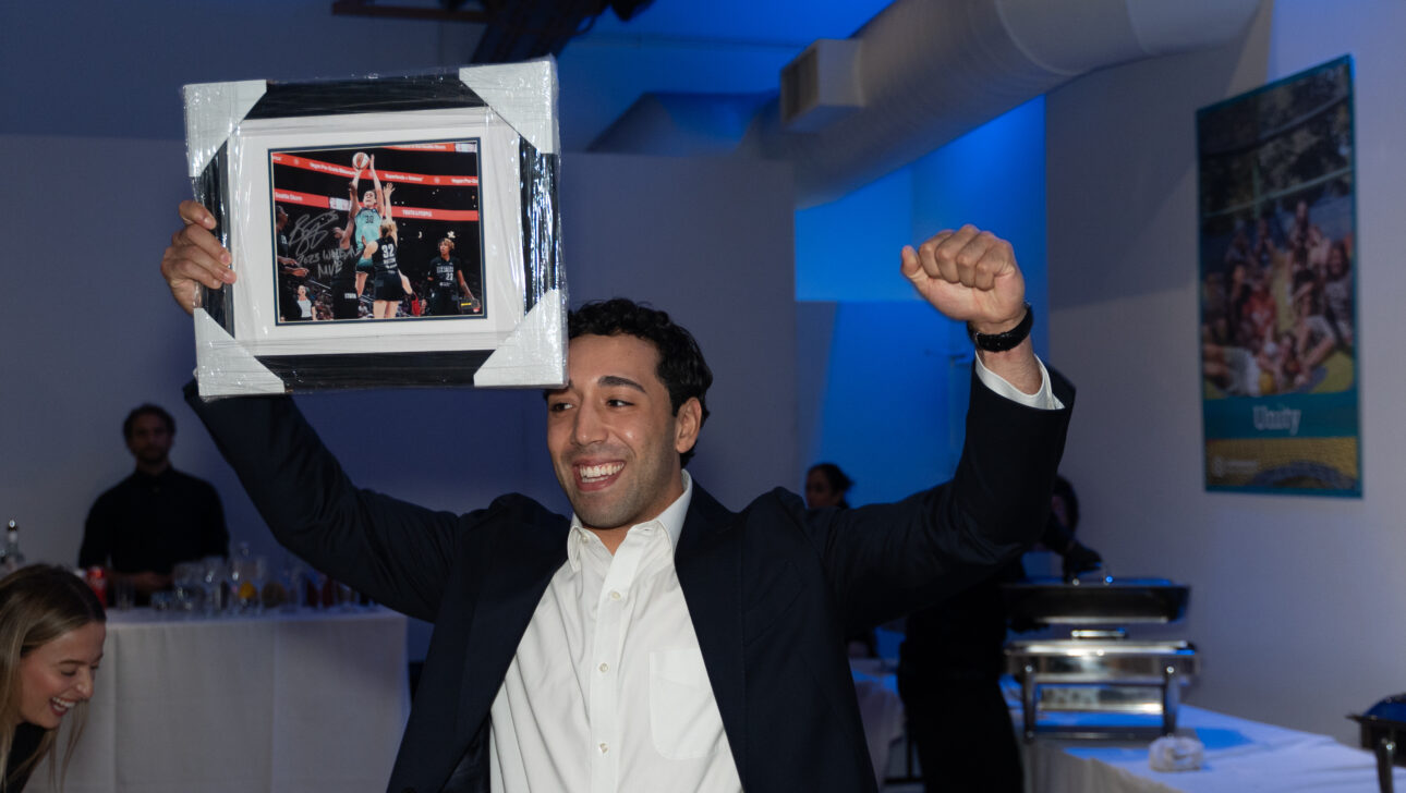 At the Commonpoint Gala, a man in a suit holds up a framed sports photograph with arms raised, smiling in a room with a buffet table and blue lighting.