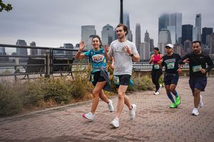Runners participate in a race on a paved path with the city skyline in the background; two runners in front wave at the camera during the TCS New York City Marathon.