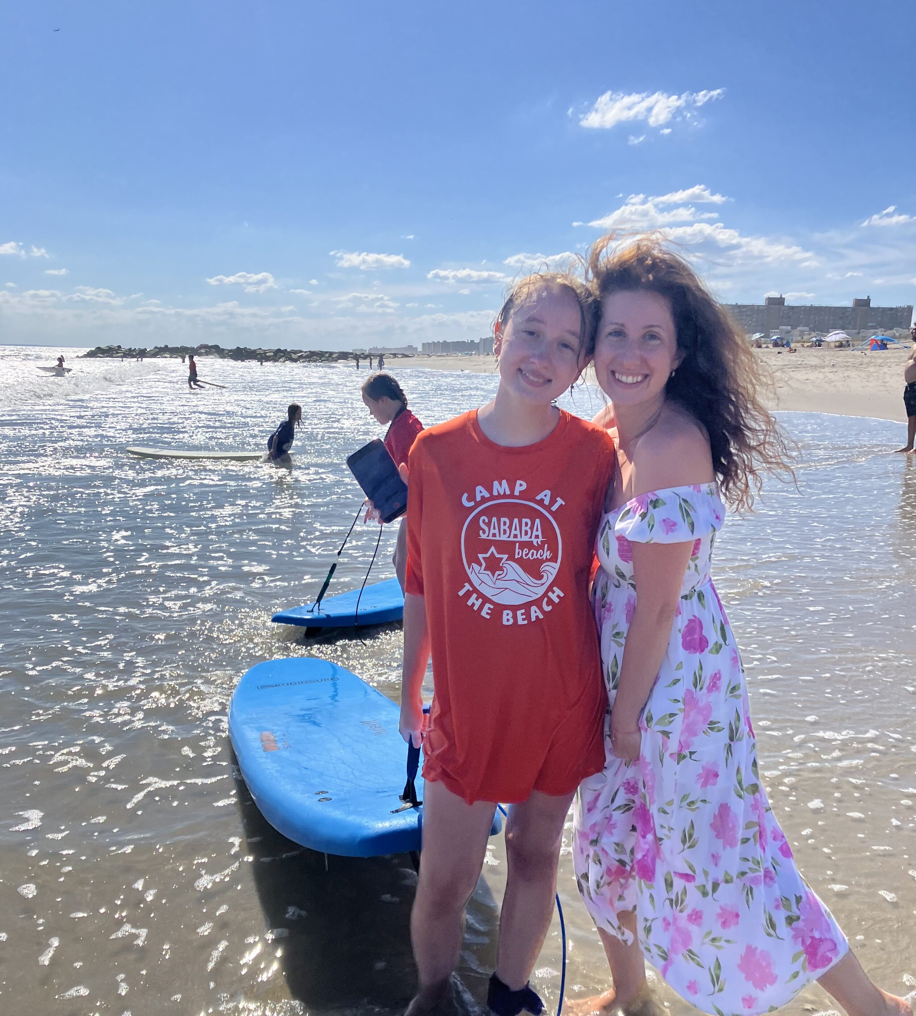 Two people stand on a beach in shallow water near blue paddleboards at Sababa Beach Camp, enjoying a vibrant summer experience, with other beachgoers and the ocean in the background.