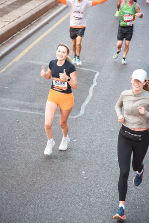 A group of marathon runners on a city street, with one woman in orange shorts giving a thumbs-up to the camera during the TCS New York City Marathon.
