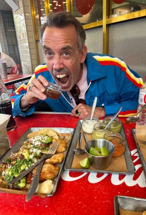 A man in a colorful jacket sits at a red table, savoring tacos and sauces after the TCS New York City Marathon, holding a small cup near his mouth as he celebrates his run.