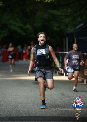 A man in athletic gear runs towards the finish line of the New York City Marathon, with other runners behind him and trees in the background. An ice cream cone logo is visible in the corner.