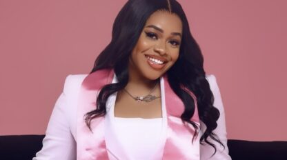 Woman in a white suit with a pink satin stole sits on a black couch, smiling, against a solid pink background.