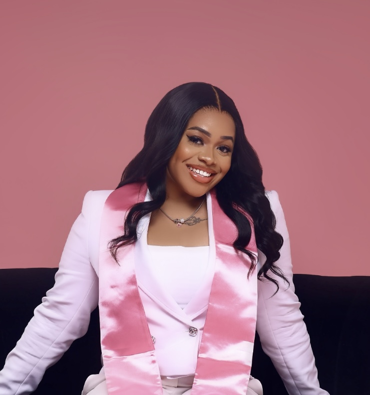 Woman in a white suit with a pink satin stole sits on a black couch, smiling, against a solid pink background.