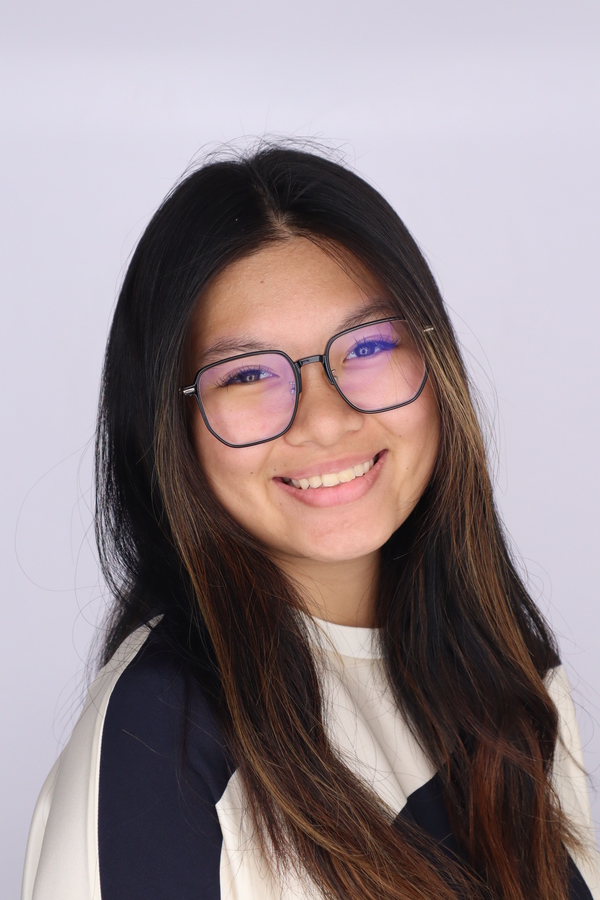 A young woman with long dark hair and glasses smiles at the camera against a plain light background. She is wearing a white and black top.