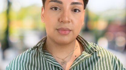 A person with short dark hair wearing a green and white striped shirt and necklace stands outdoors with a blurred background.
