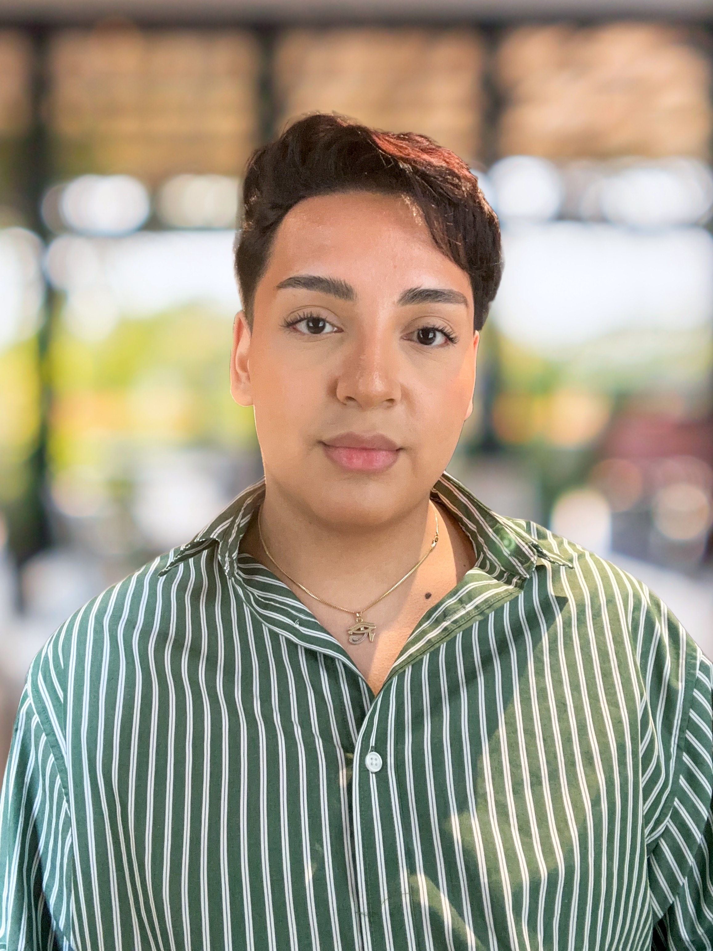 A person with short dark hair wearing a green and white striped shirt and necklace stands outdoors with a blurred background.