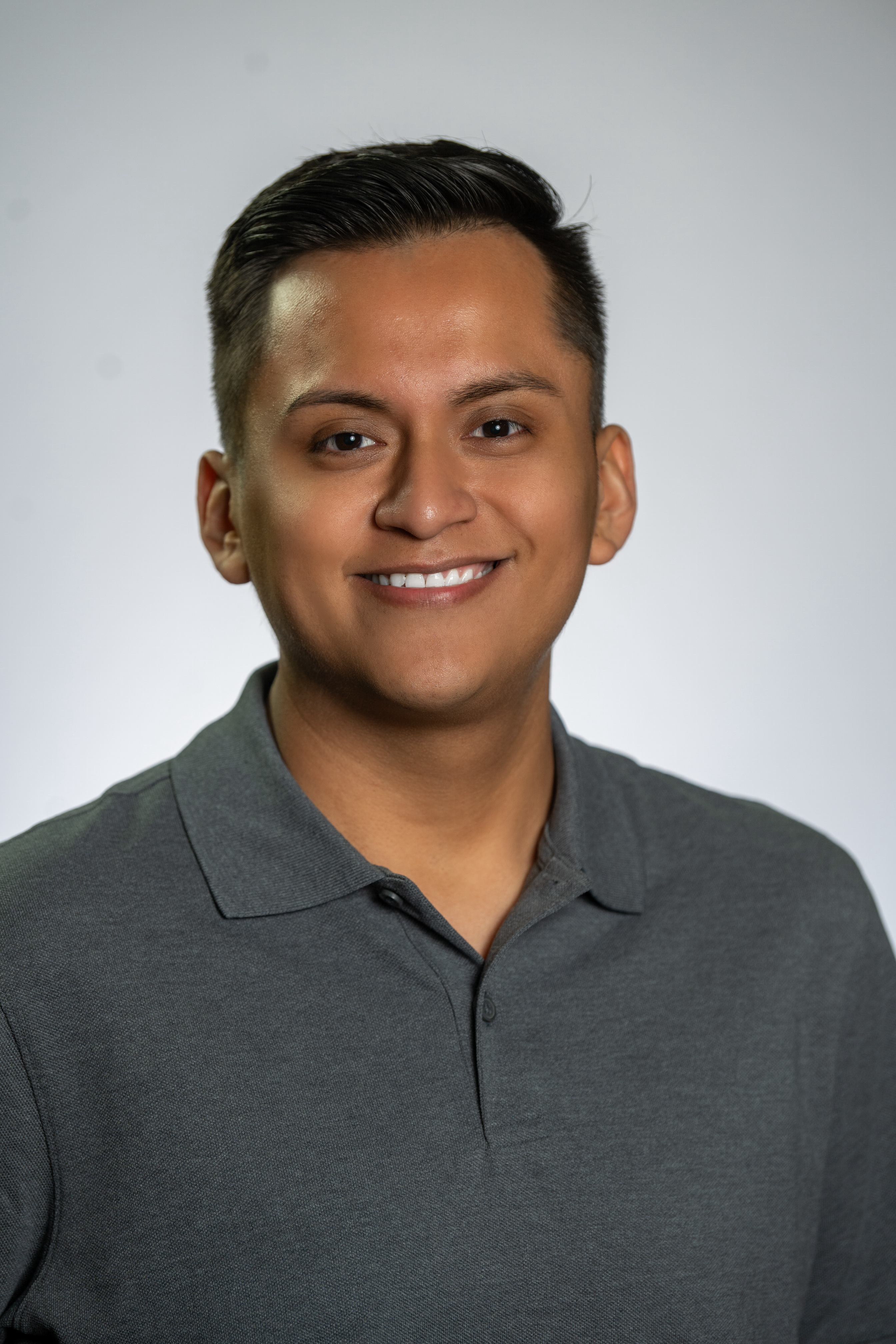 A man with short dark hair wearing a gray polo shirt smiles at the camera against a plain light background.