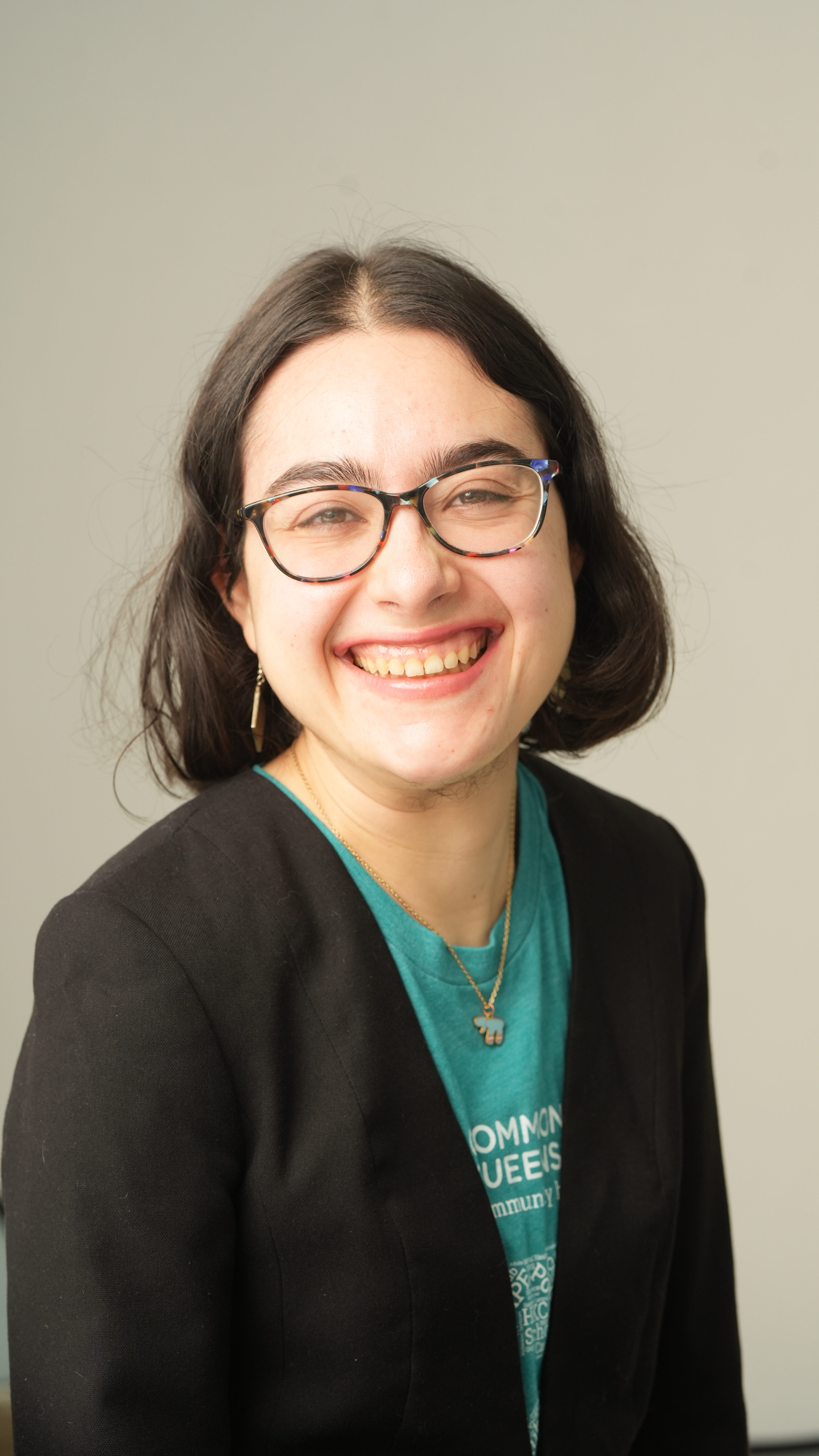 A person with shoulder-length dark hair, glasses, and a black blazer over a teal shirt smiles at the camera against a plain light background.