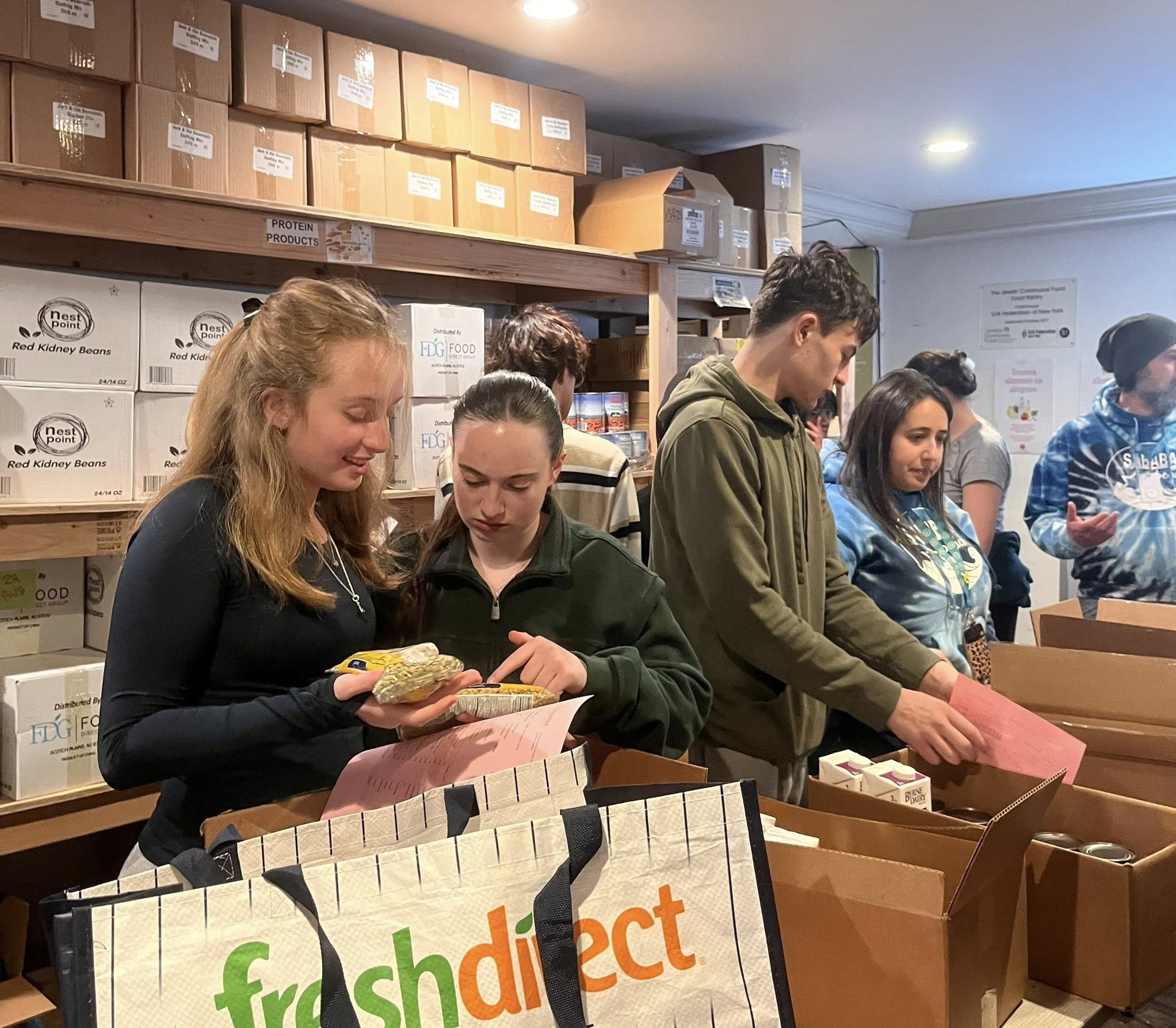 Several people pack food items into boxes in a room lined with shelves holding other boxes and supplies. A reusable FreshDirect bag sits in the foreground.