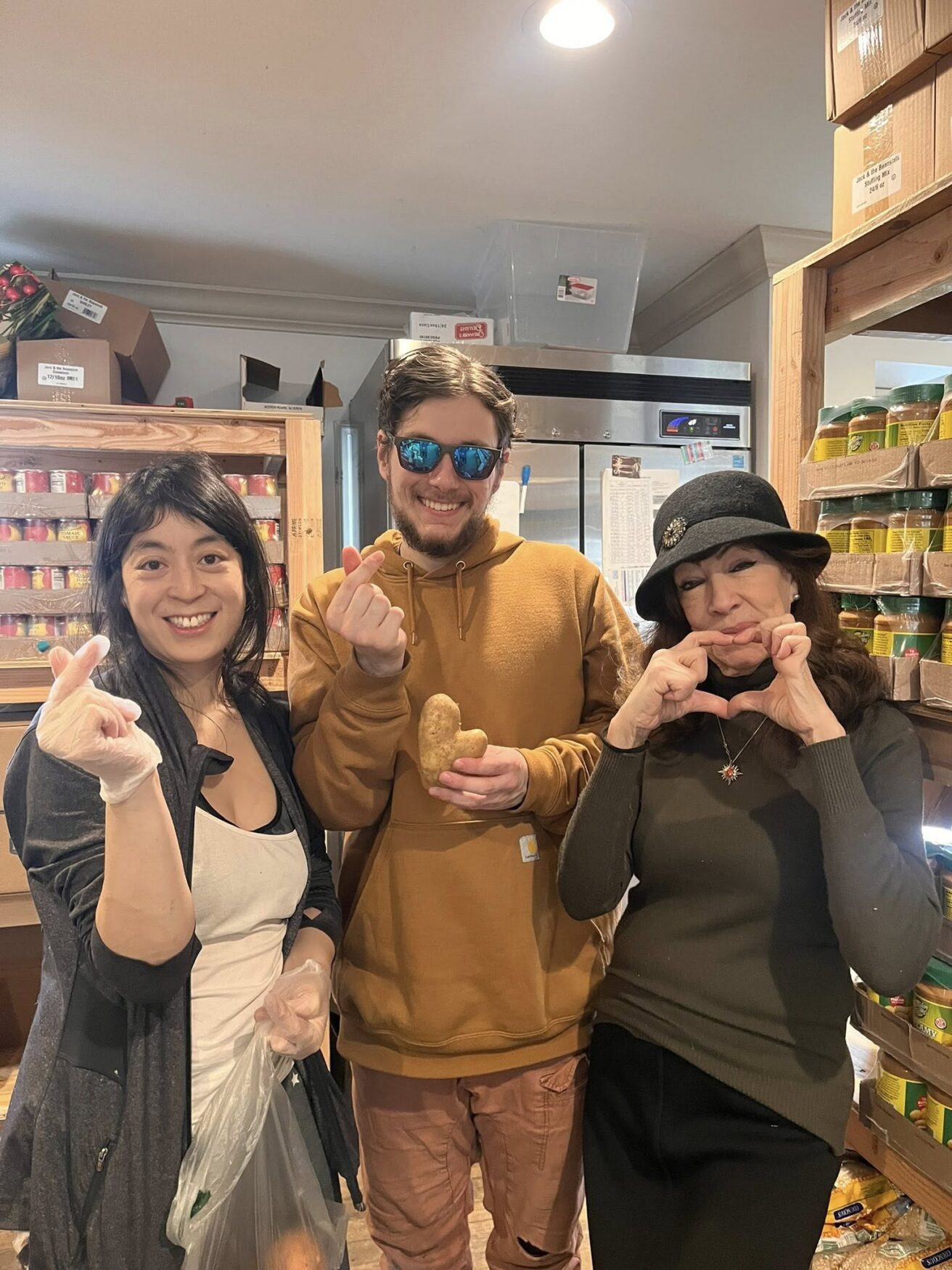 Three people standing in a food pantry, smiling at the camera. The man in the middle holds a potato; shelves with canned goods are visible in the background.
