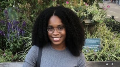 Michelle Nairne, a woman with curly hair and glasses, sits on a bench in a garden, smiling at the camera. Green plants and flowers add charm to the background.