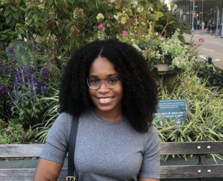 Michelle Nairne, a woman with curly hair and glasses, sits on a bench in a garden, smiling at the camera. Green plants and flowers add charm to the background.