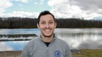 Jeff Horowitz stands smiling by a lake in a gray zip-up jacket, with a dock and trees in the background beneath a partly cloudy sky.