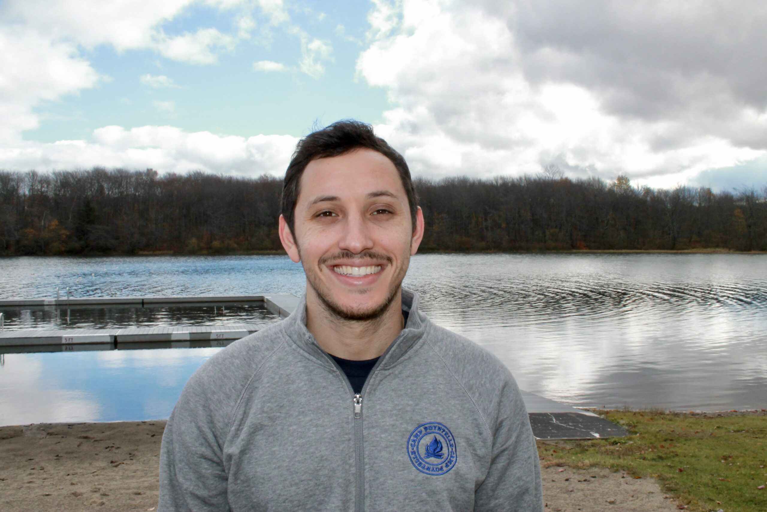 Jeff Horowitz stands smiling by a lake in a gray zip-up jacket, with a dock and trees in the background beneath a partly cloudy sky.