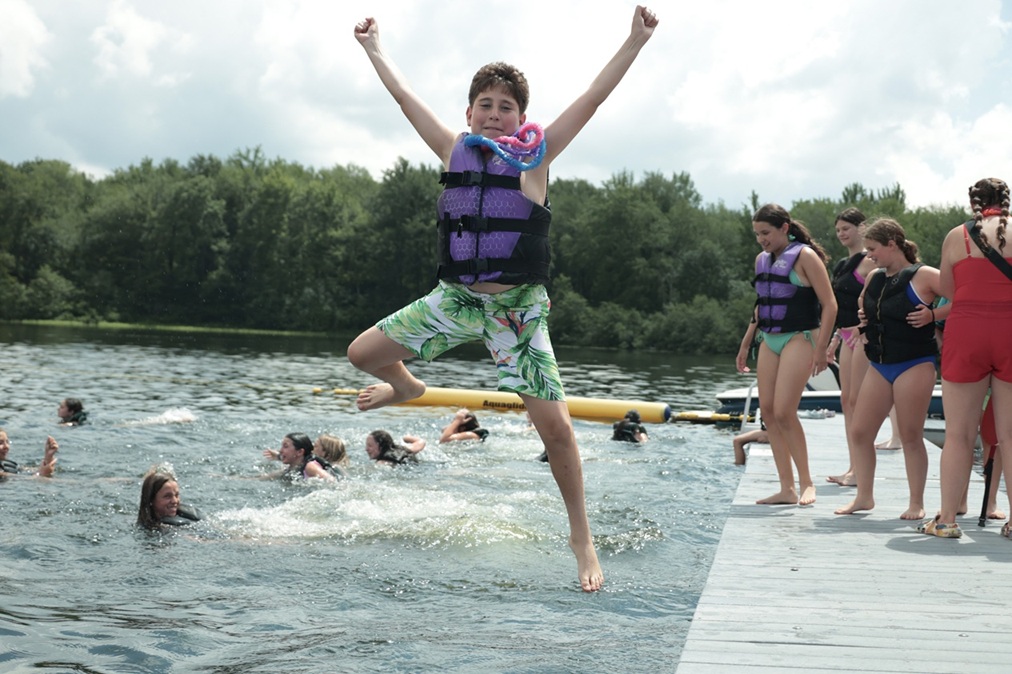 A boy in a life jacket and swim trunks jumps off a dock into a lake, with other children swimming and standing nearby.