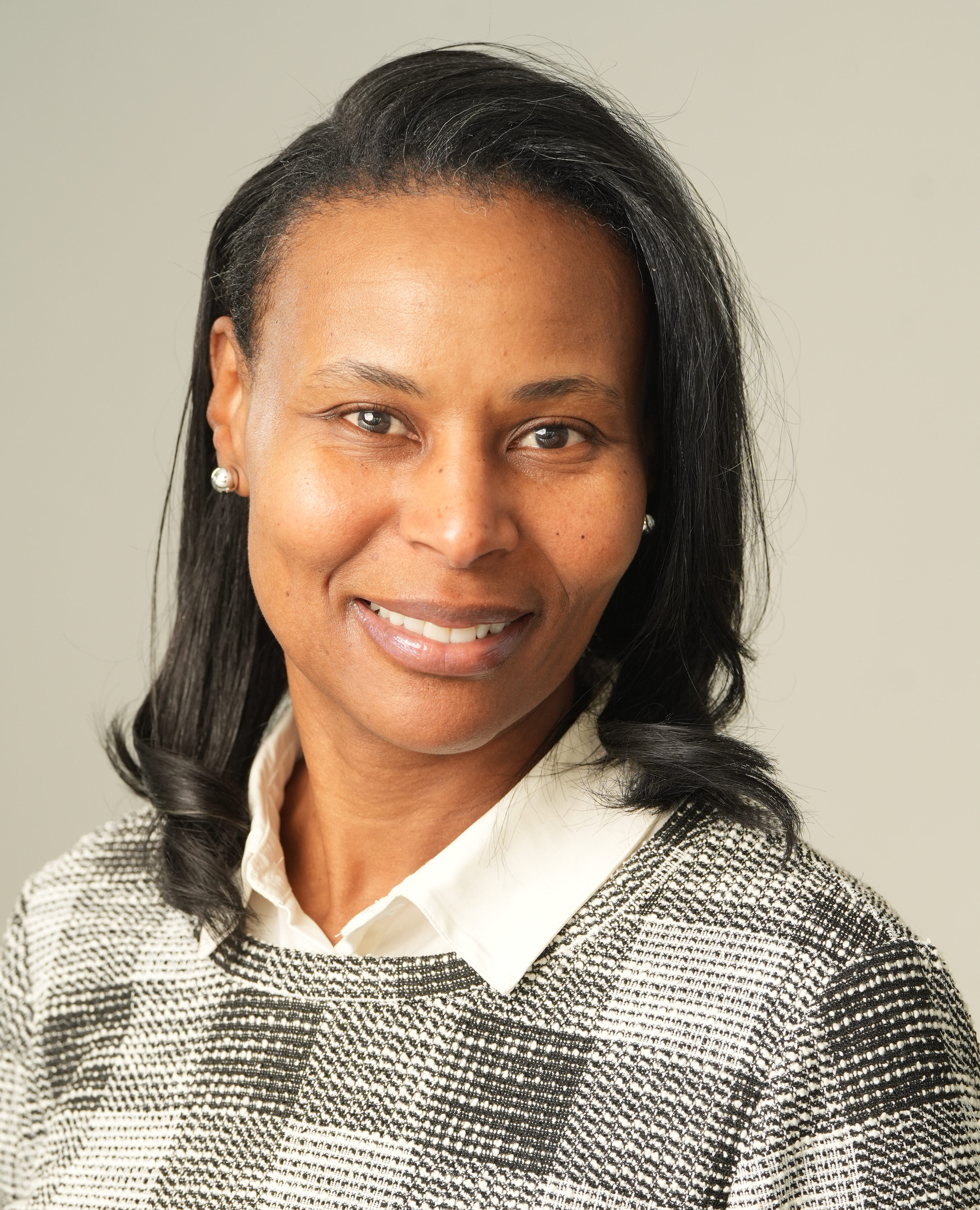 Sabrina Perrin, with shoulder-length black hair, smiles at the camera in a black-and-white checkered sweater over a white collared shirt, set against a neutral background.