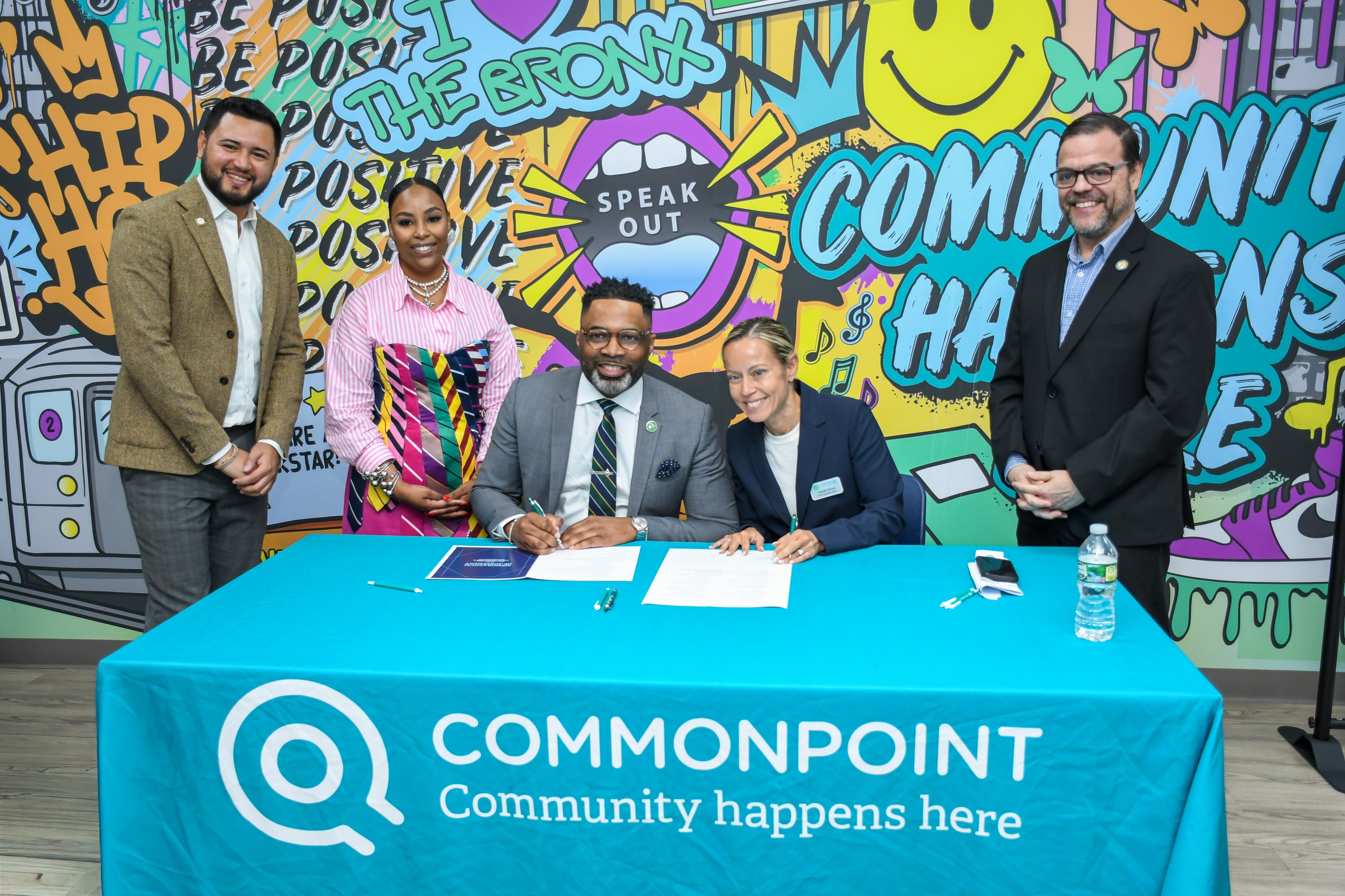Five people pose at a table with “Commonpoint Community happens here” printed on the tablecloth. Two are seated, signing documents related to workforce opportunities, while three stand behind them against a colorful mural background.