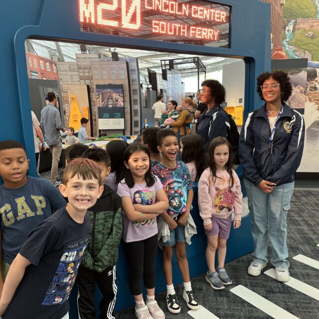 A group of children and an adult pose and smile together inside an indoor exhibit with a digital sign reading "Lincoln Center South Ferry.