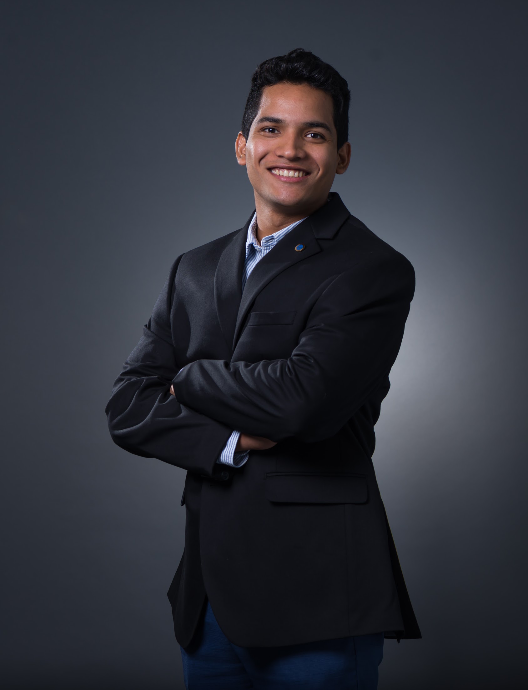 A man in a dark suit jacket stands with arms crossed and smiles at the camera against a plain dark background.