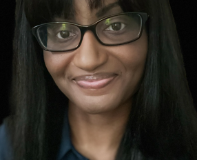 A person with long straight hair, wearing glasses and a dark shirt, smiles softly at the camera against a dark background.