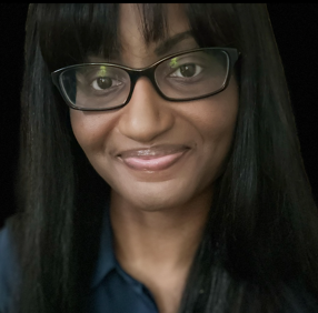 A person with long straight hair, wearing glasses and a dark shirt, smiles softly at the camera against a dark background.