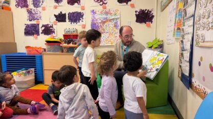 A teacher reads a picture book to a group of young children in a colorful classroom decorated with children's artwork during the COMMONPOINT CHALLENGE.