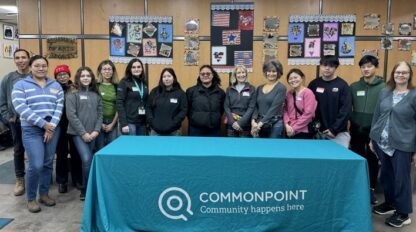 A group of people stand behind a table with a teal Commonpoint tablecloth, ready to take on the Commonpoint challenge in a room decorated with artwork and crafts on the wall.