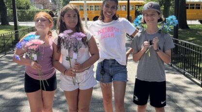 Four children stand outdoors holding colorful flowers, with a school bus and flagpole in the background on a sunny day, ready to take on the COMMONPOINT CHALLENGE.