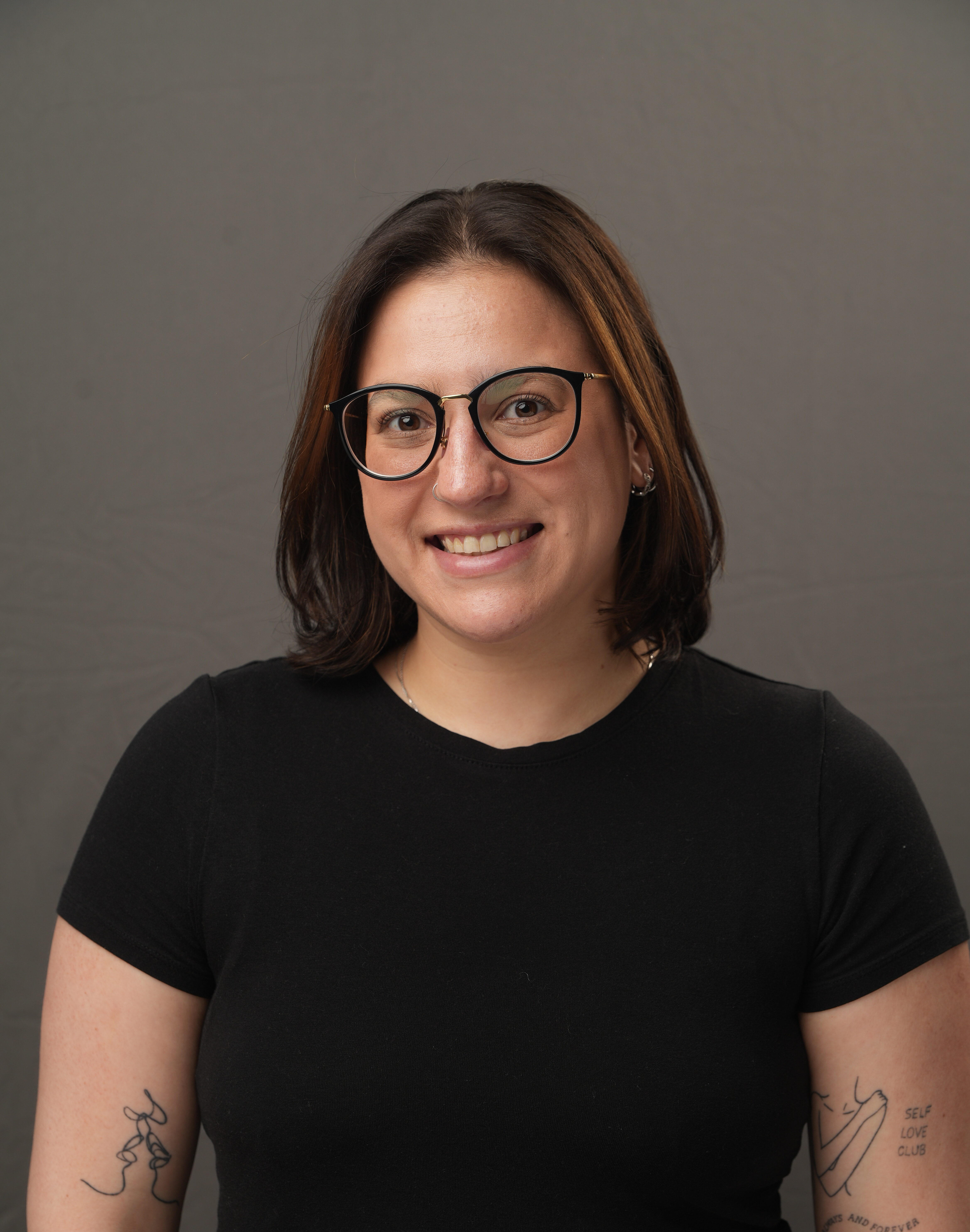 Stephanie Silberman, wearing glasses and a black t-shirt, smiles at the camera in front of a plain gray background. Tattoos are visible on both arms.