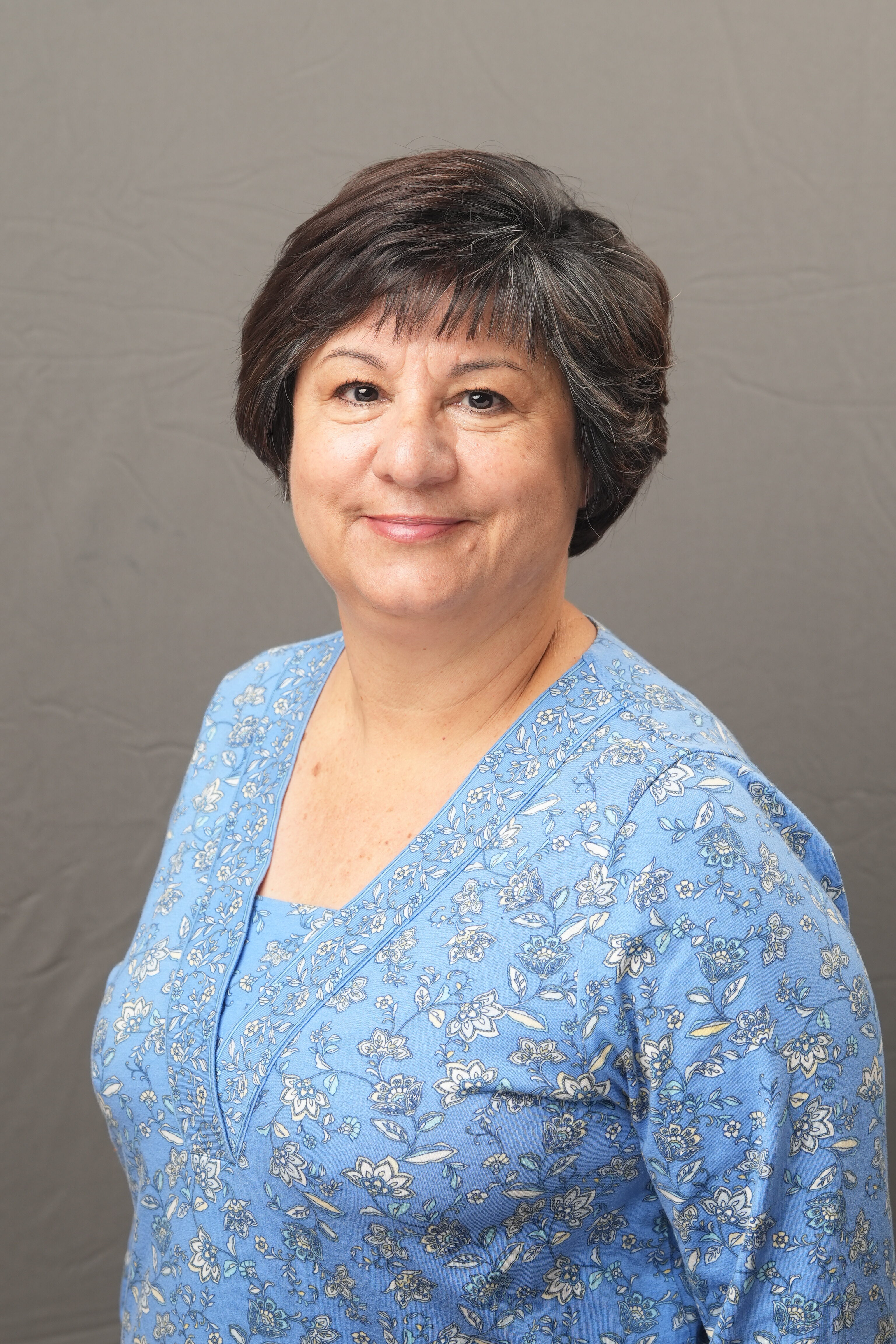 A woman with short dark hair wearing a blue floral blouse stands against a plain gray background, facing slightly to the side and smiling gently.
