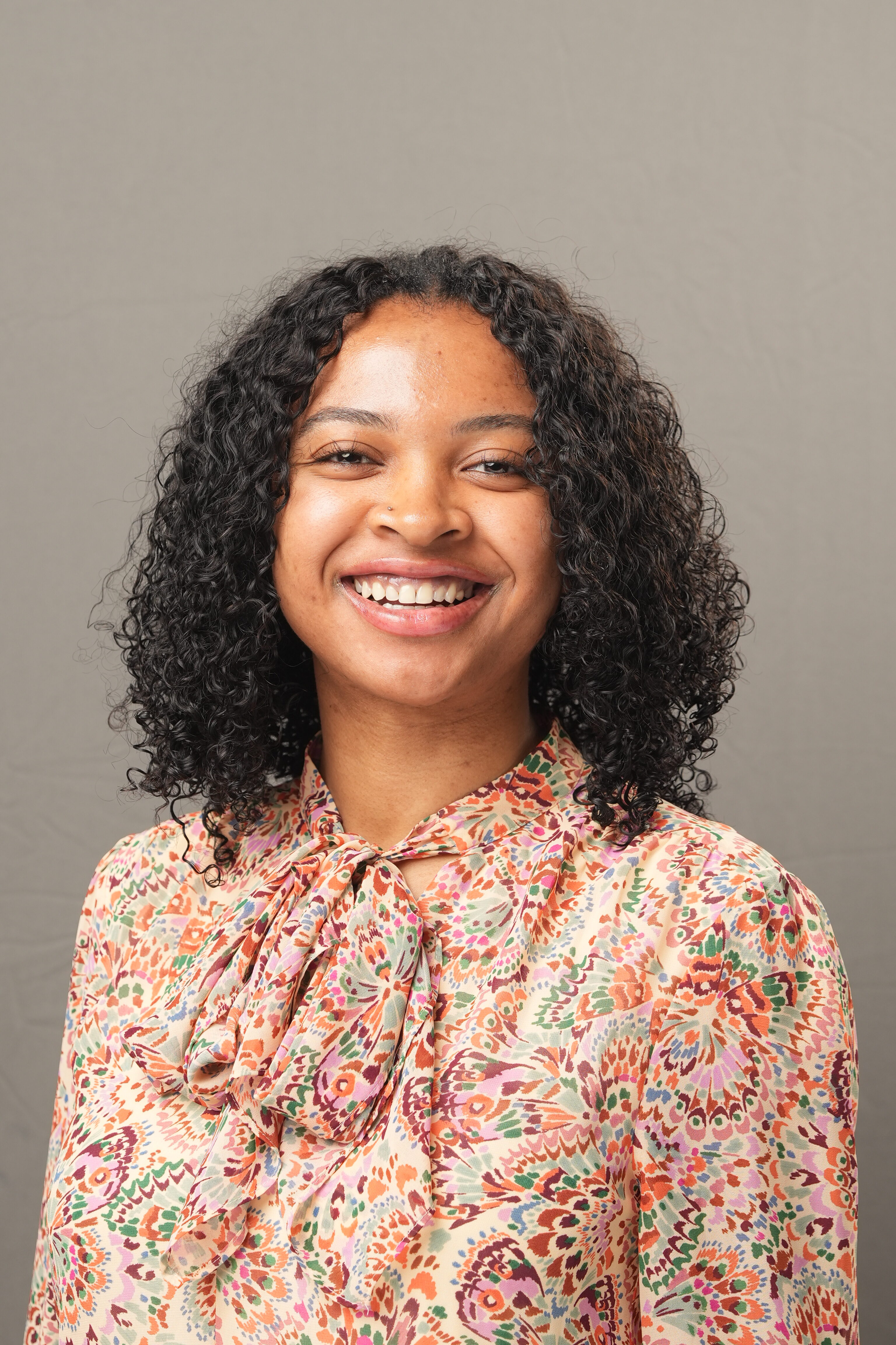 A woman with curly hair smiles at the camera, wearing a colorful patterned blouse with a bow at the neckline, against a plain background.