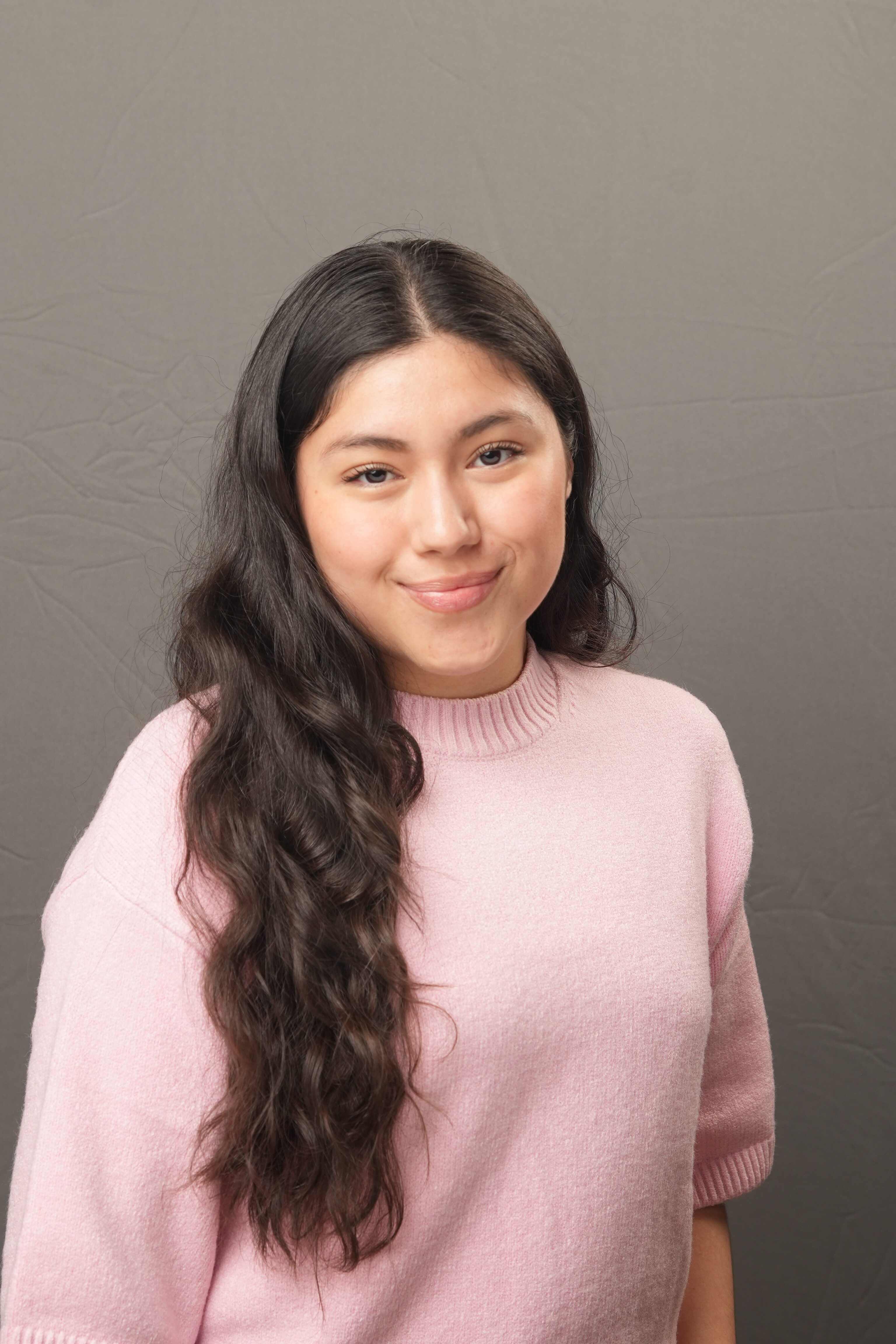 A young woman with long, wavy dark hair wearing a light pink sweater poses in front of a plain gray background.
