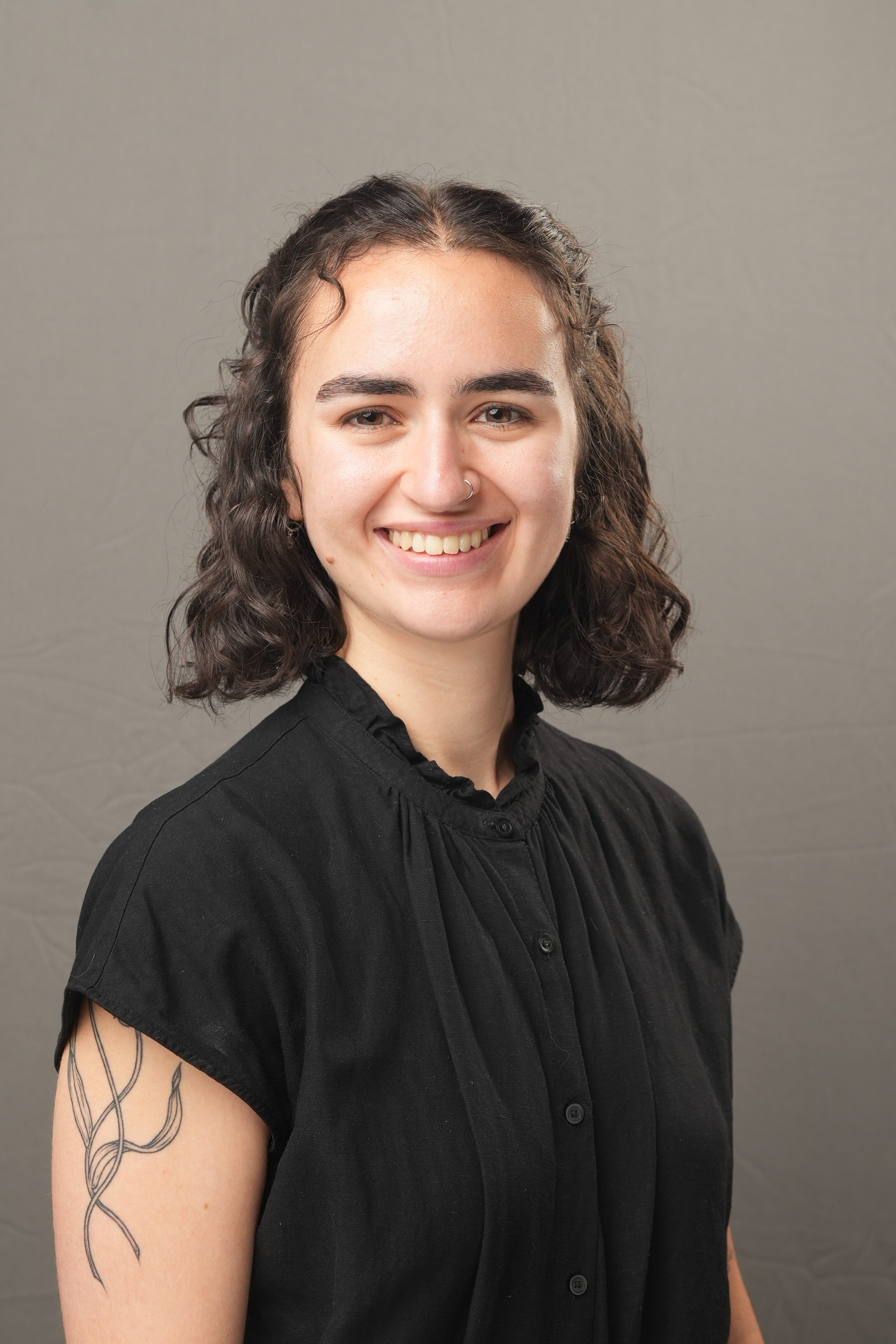 A person with short curly hair wearing a black shirt, smiling at the camera. They have a tattoo on their upper arm and a nose ring. The background is plain and neutral.