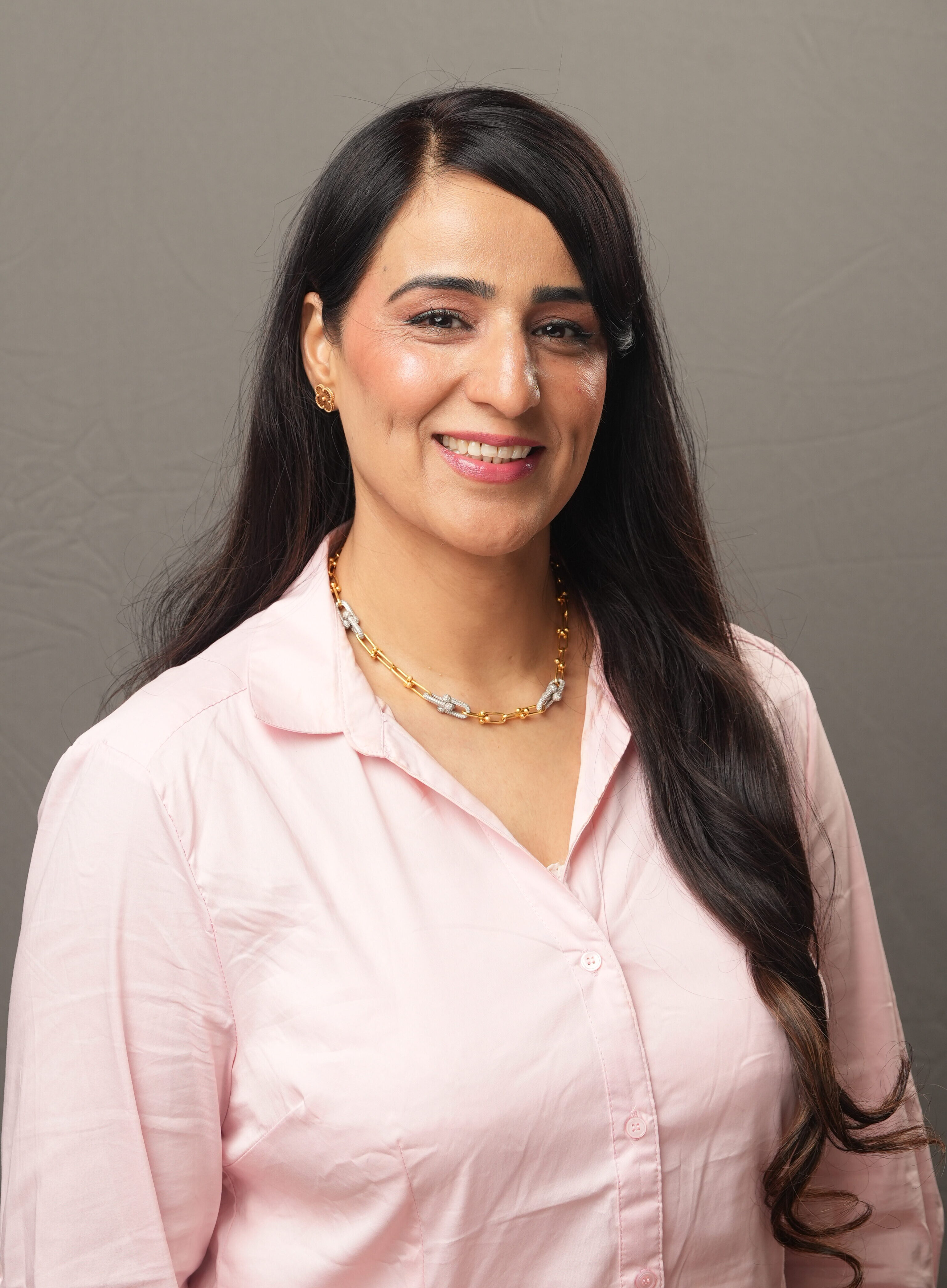 A woman with long dark hair wearing a light pink button-up shirt and a gold necklace smiles against a plain gray background.