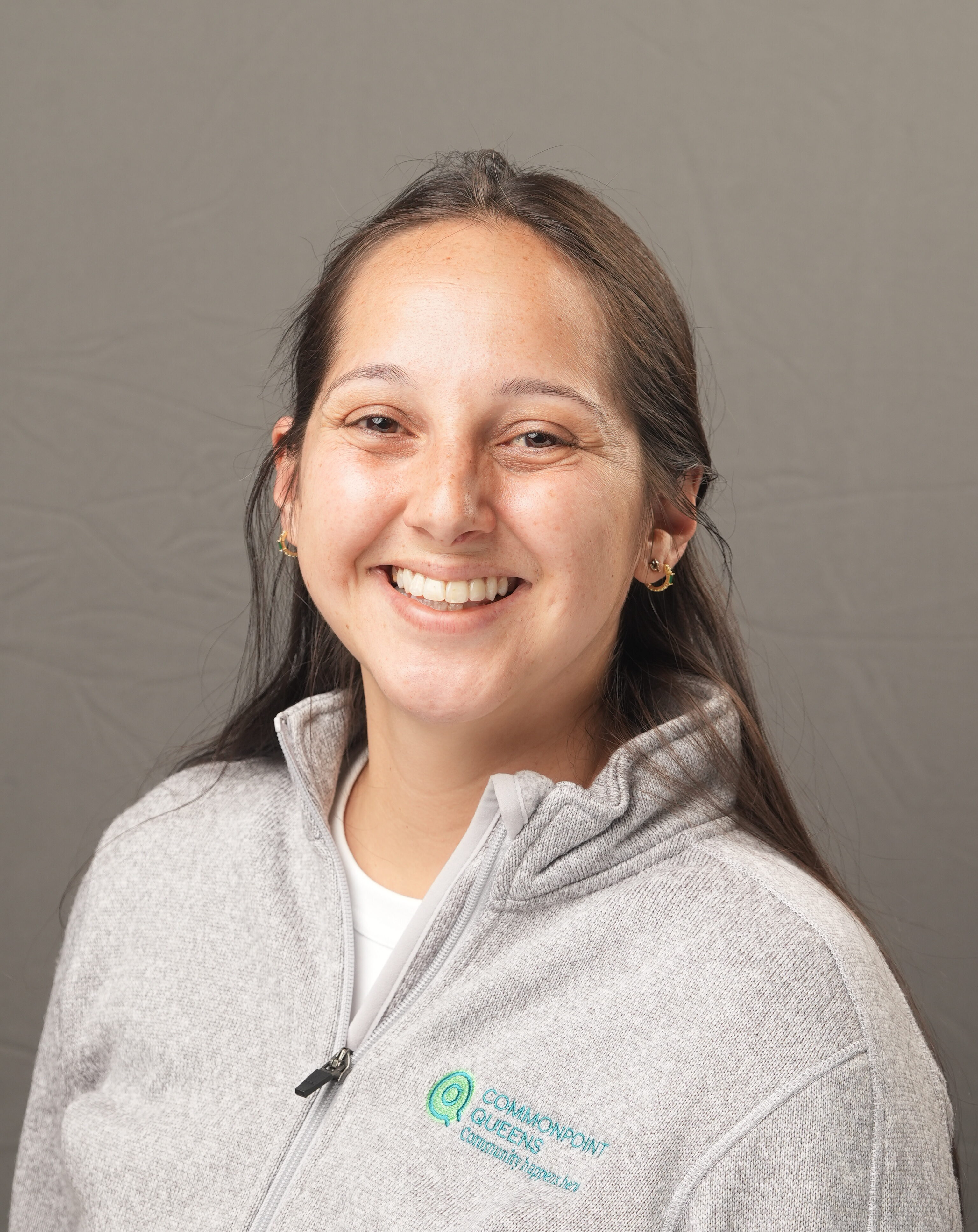 A woman with long brown hair smiles at the camera, wearing a light gray zip-up jacket with a "Commonpoint Queens" logo on the left side. The background is plain gray.