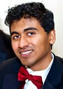 A man in formal attire with a red bow tie and dark suit smiles at the camera against a neutral background.