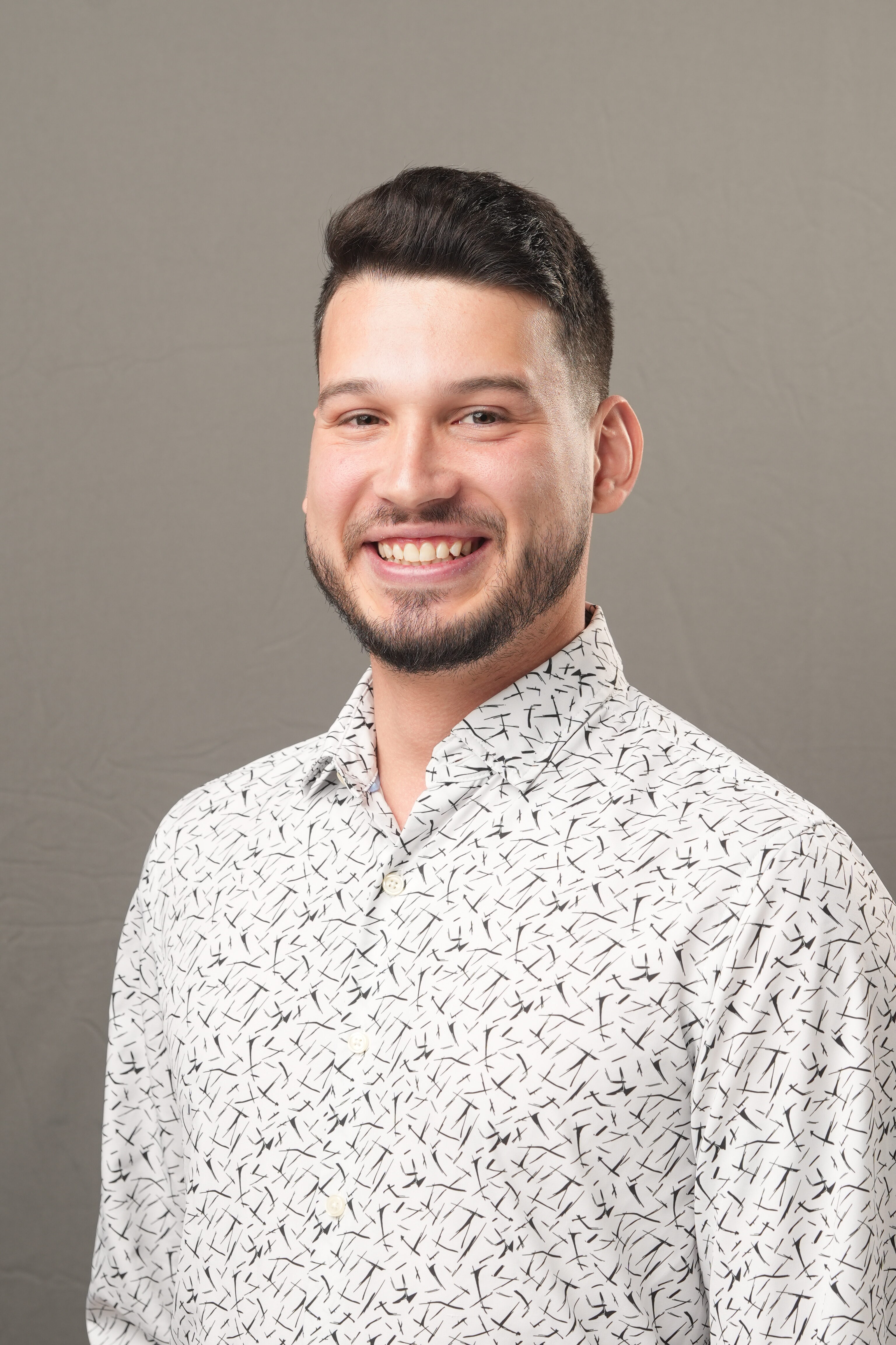 A man with short dark hair and a beard smiles at the camera, wearing a white Carras button-up shirt with a black abstract pattern, in front of a plain gray background.